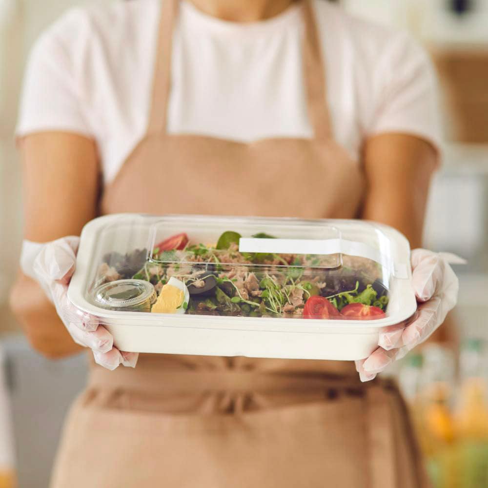 Operatore mensa con guanti che porge una vaschetta compostabile con coperchio trasparente, contenente un'insalata fresca con ingredienti naturali.