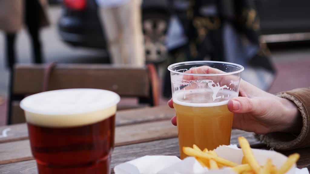 Bicchiere compostabile con birra chiara in primo piano e birra ambrata su tavolo all’aperto con patatine fritte