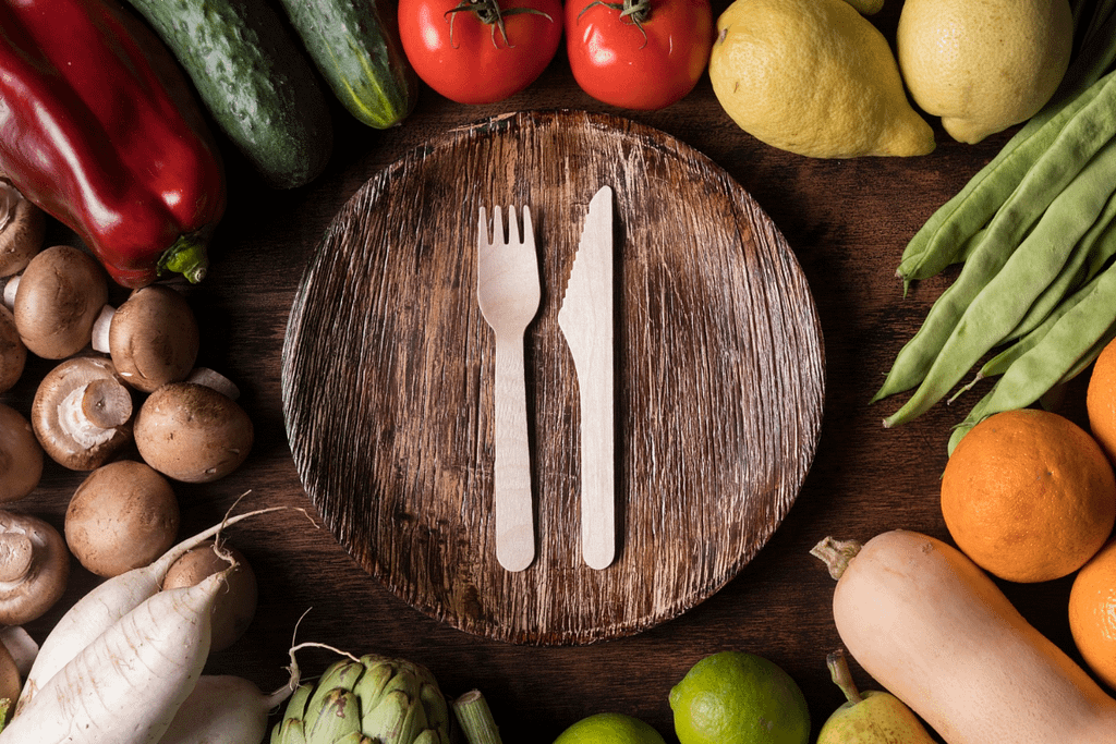 Piatto in foglia di palma con posate compostabili in legno su tavolo in legno circondato da verdure fresche — soluzioni sostenibili Ekoe per ristorazione ed eventi.