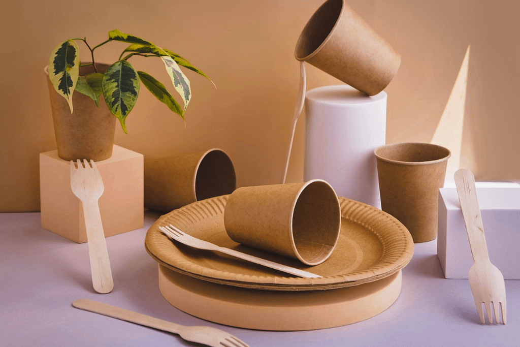 Stoviglie compostabili color avana: piatti, bicchieri e posate in legno, con una pianta decorativa su sfondo neutro