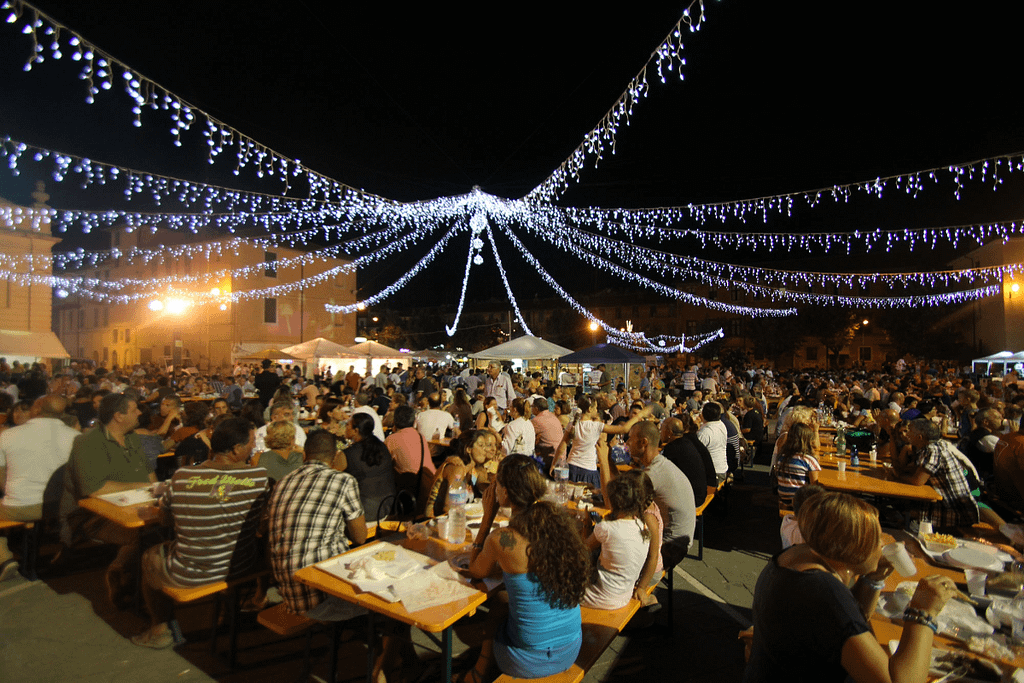 Sagra di paese serale con tavoli all’aperto, persone e luminarie festive